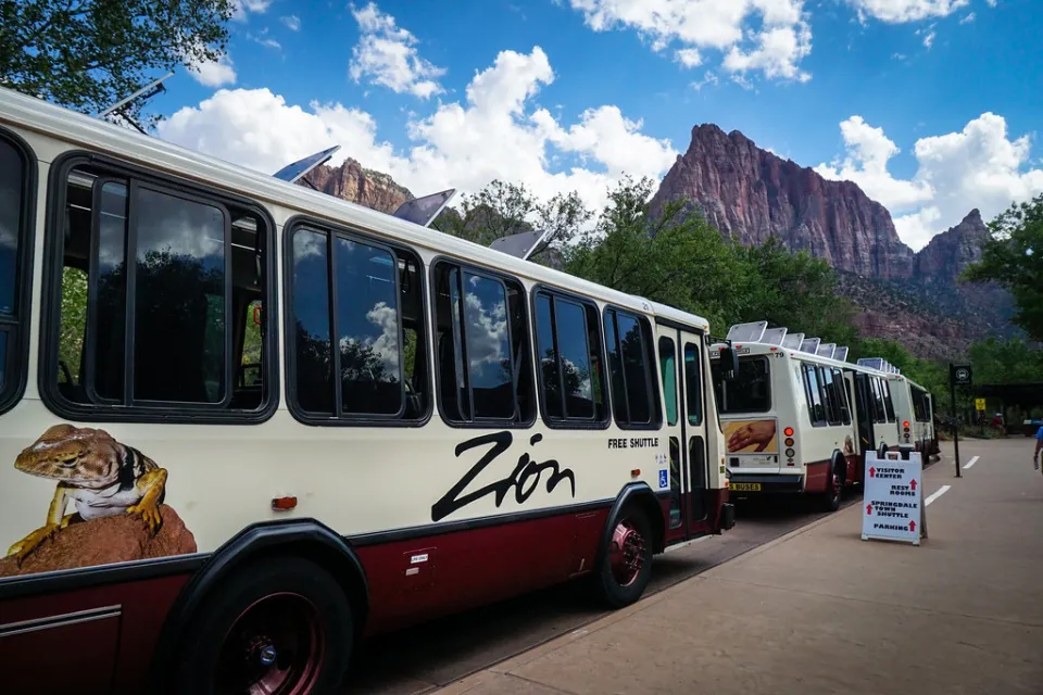 White park shuttles with large Zion logo on side and red rock cliffs in background in Zion National Park.